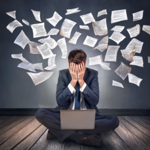 man in a suit hands over his face, papers flying in the air - laptop in the front. stress anxiety