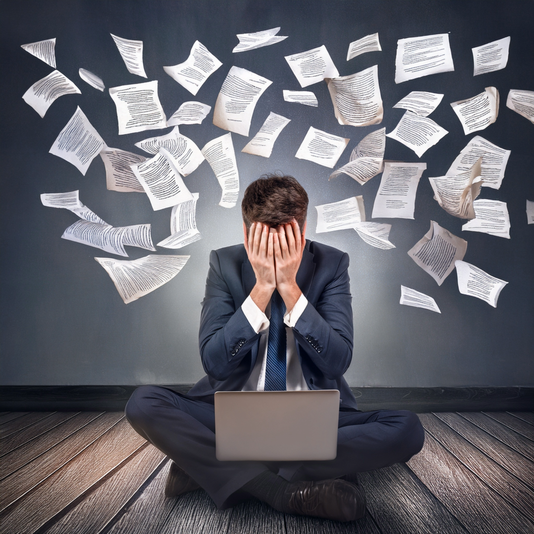 man in a suit hands over his face, papers flying in the air - laptop in the front. stress anxiety
